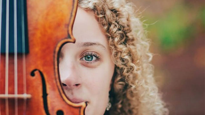 Half of a young woman's face peaks out from behind a violin, which is in the foreground.