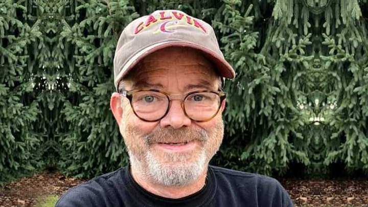 A man with glasses and facial hair wearing a Calvin ballcap smiles with evergreen trees behind him.
