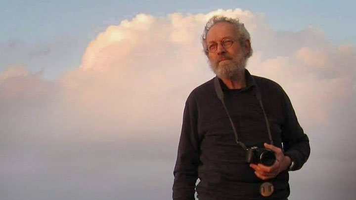 A man with a camera stares off into the distance with a cloud behind him.