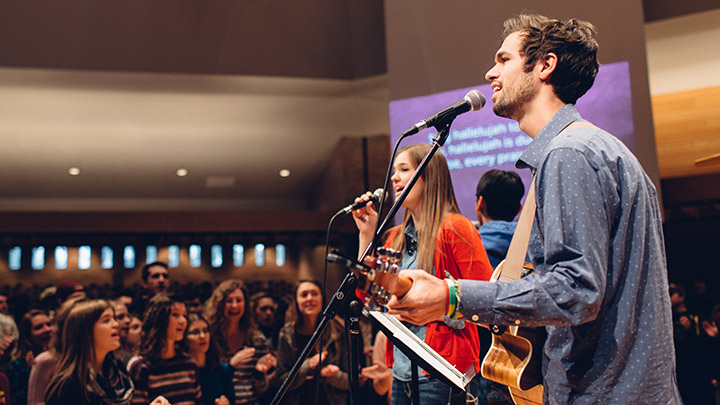 Students play guitar and sing in chapel for audience of fellow students.