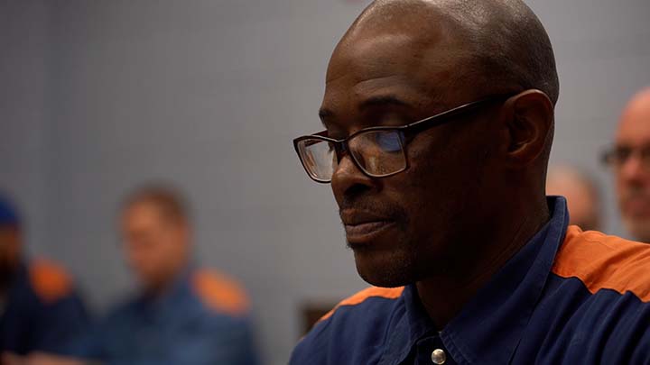 A closeup of a bald, middle-aged African American male wearing glasses and a blue shirt.