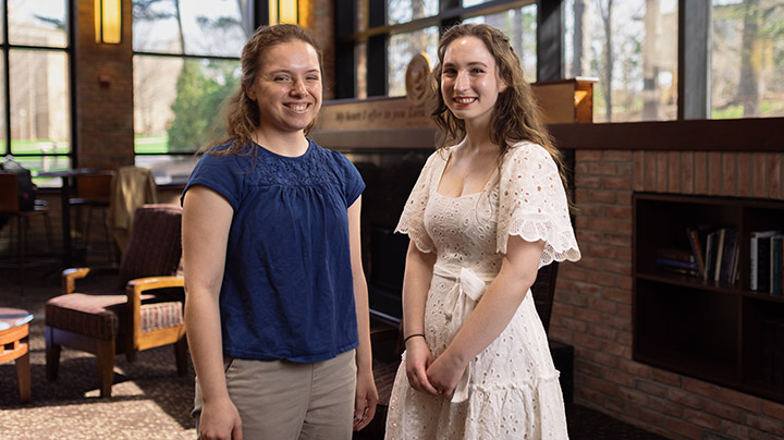 Graduating seniors Sarah Gibes and Michelle Merritt stand next to each other indoors.