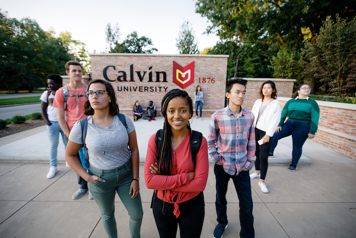 A group of students stands in a v formation in front of the Calvin University entrance sign.