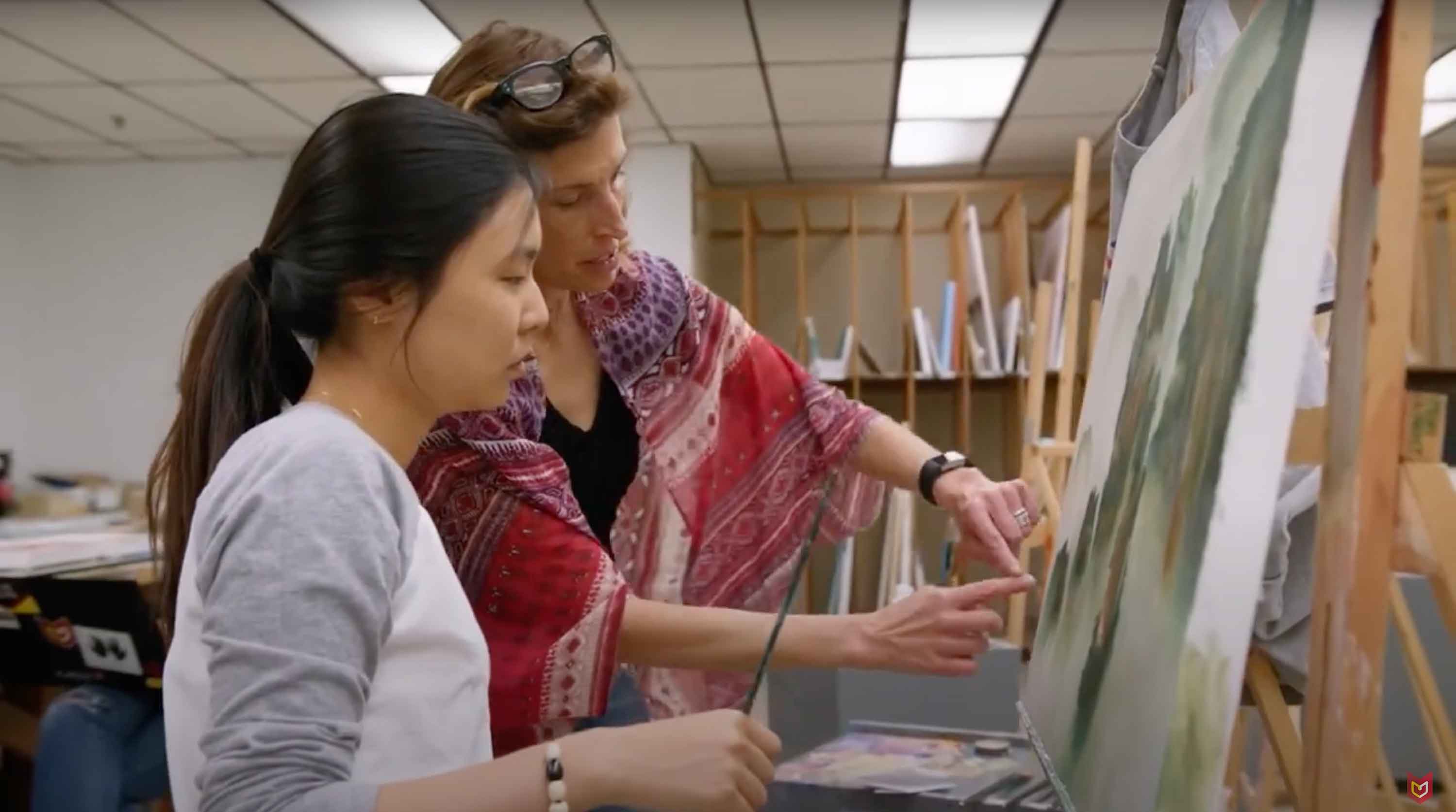 A student stands at her canvas with a paint brush, while a professor stands by and points to the canvas.