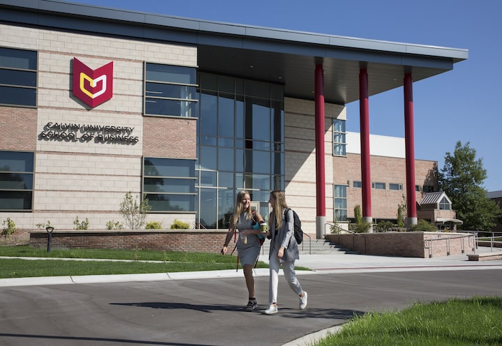 Two students walk outside in front of the Calvin University School of Business, a building with the Calvin logo, and three prominent red pillars.