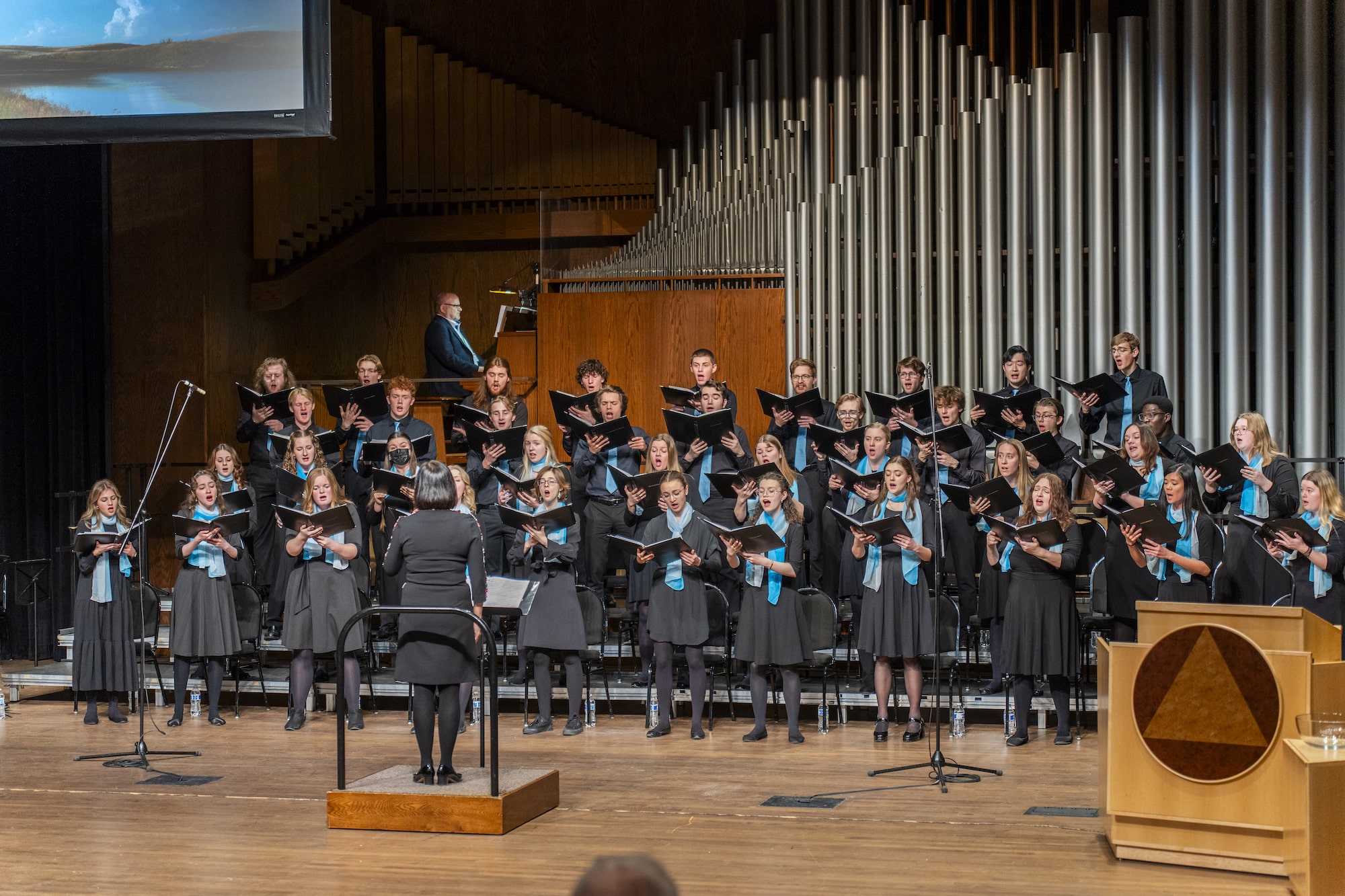 A women's choir ensemble stands in all black on risers.