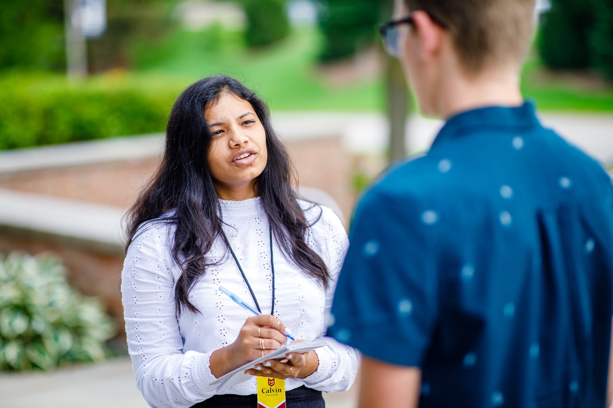 A student journalist wearing a press badge interviews someone while taking notes on a note pad.