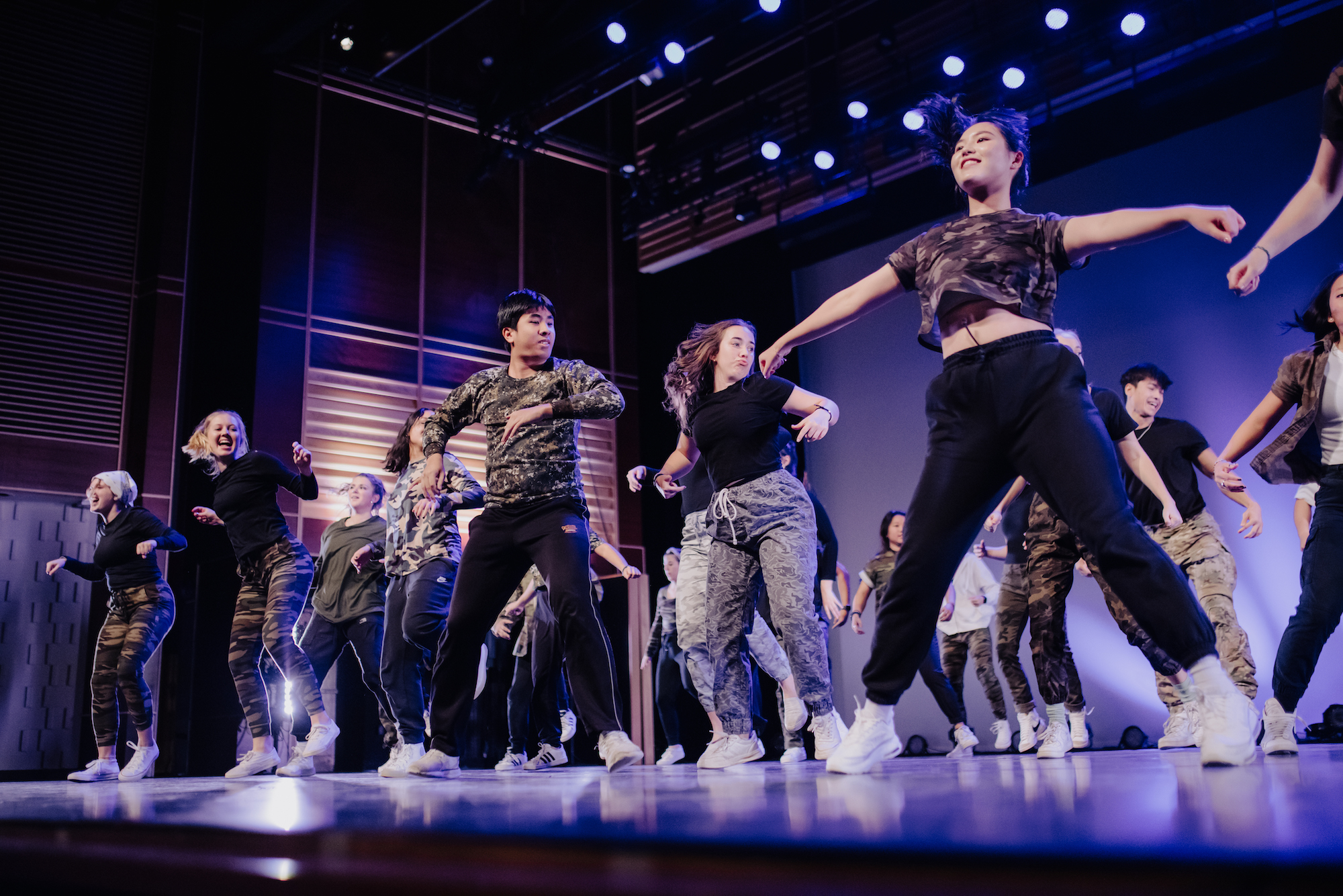 A group of male and female dancers, wearing black, white, and camo, dance in formation on the CFAC stage.