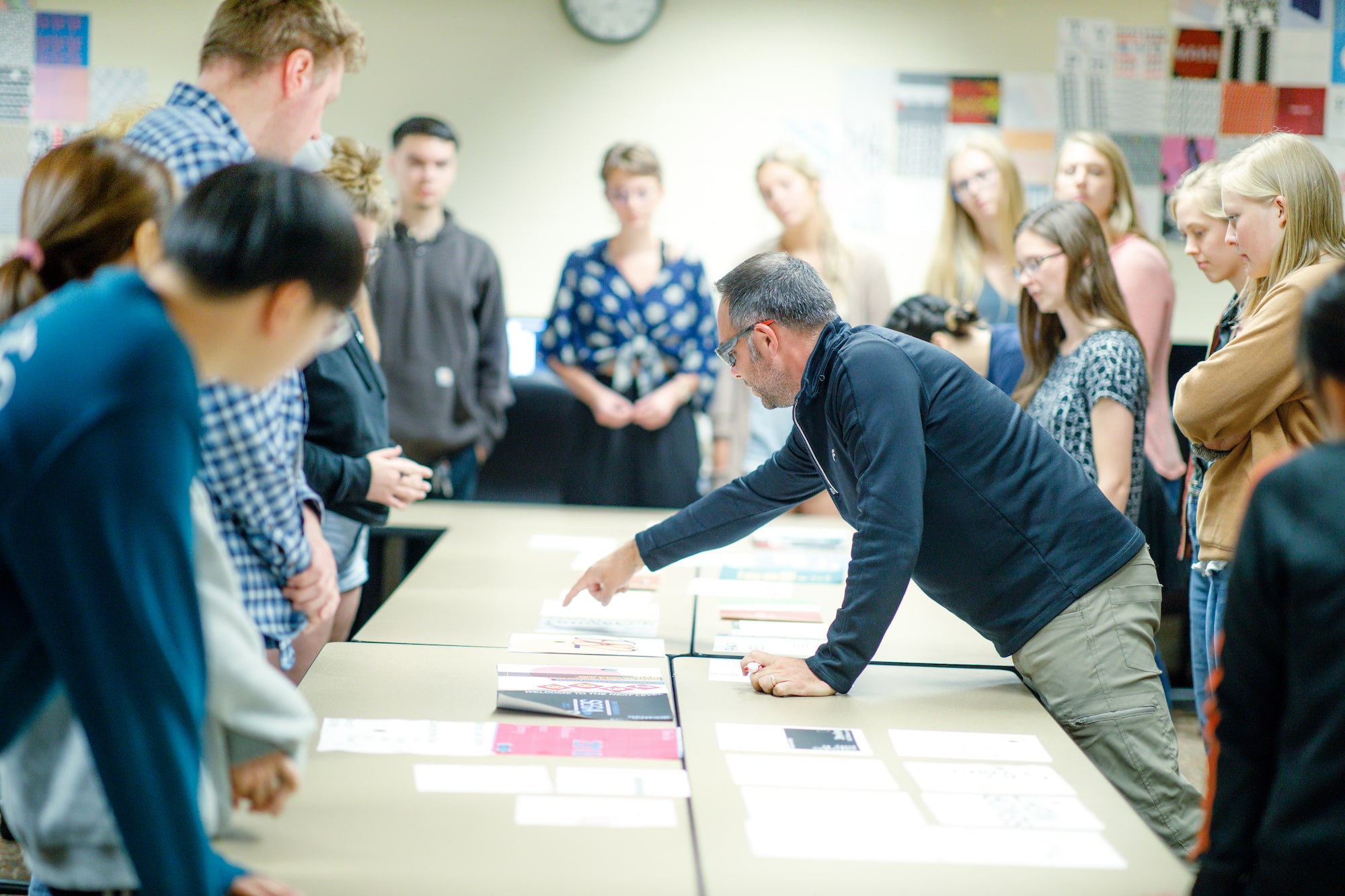 A class of students stands around tables with graphic design pieces, while a professor points at one while he teaches.