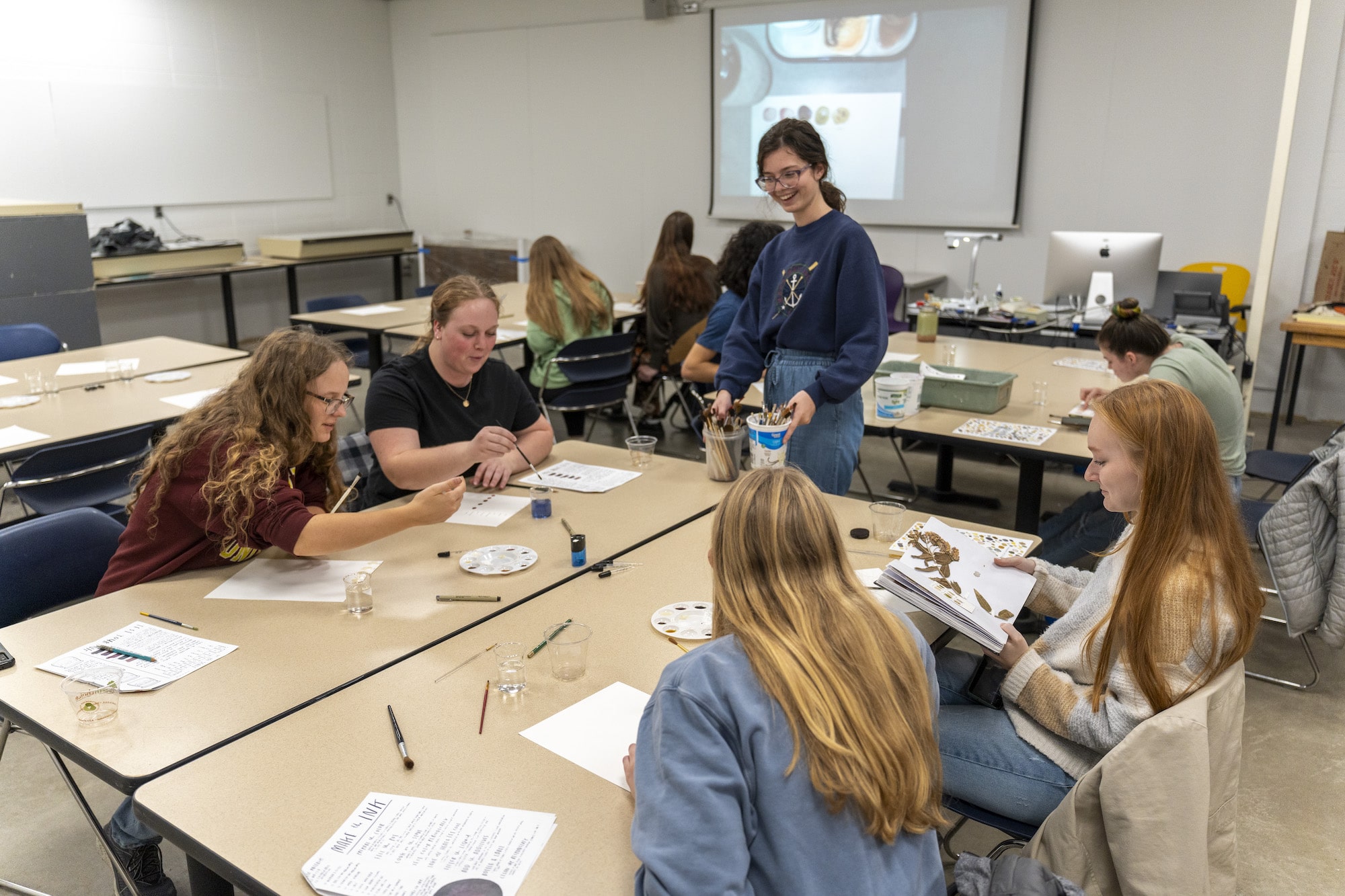 A group of students work on art projects at a table with a professor.