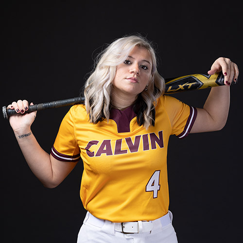 Hannah Biddlecome in her Calvin University softball uniform holding a bat behind her head across her shoulders with both hands.