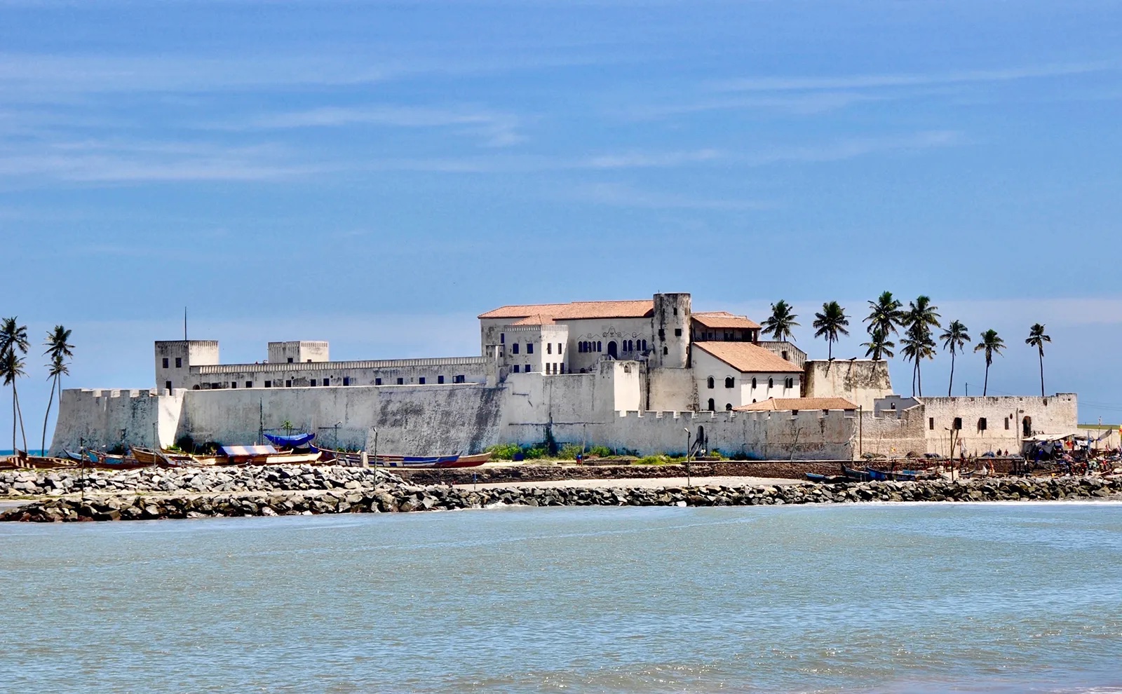 Elmina Castle under a blue sky, surrounded by palm trees.