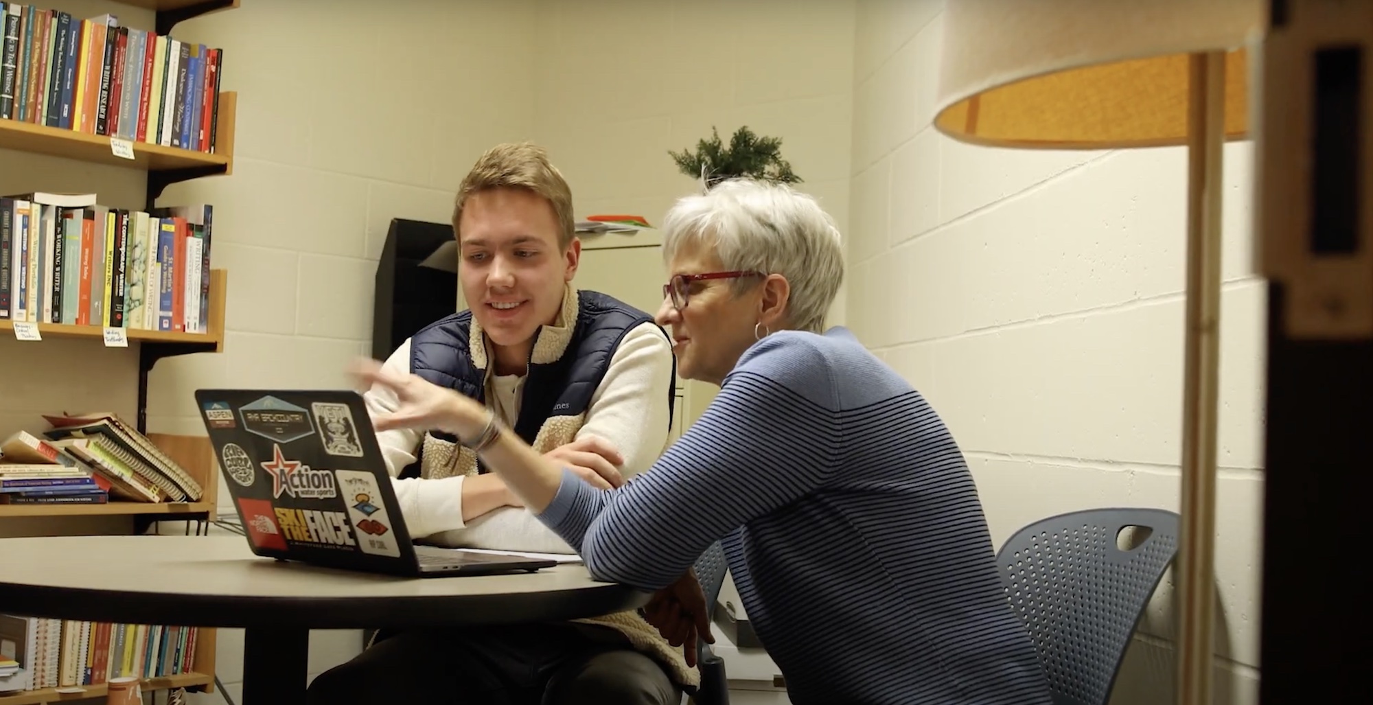 An English student and professor discuss a project on a laptop at a table in the professor's office, with plants and a bookshelf behind.