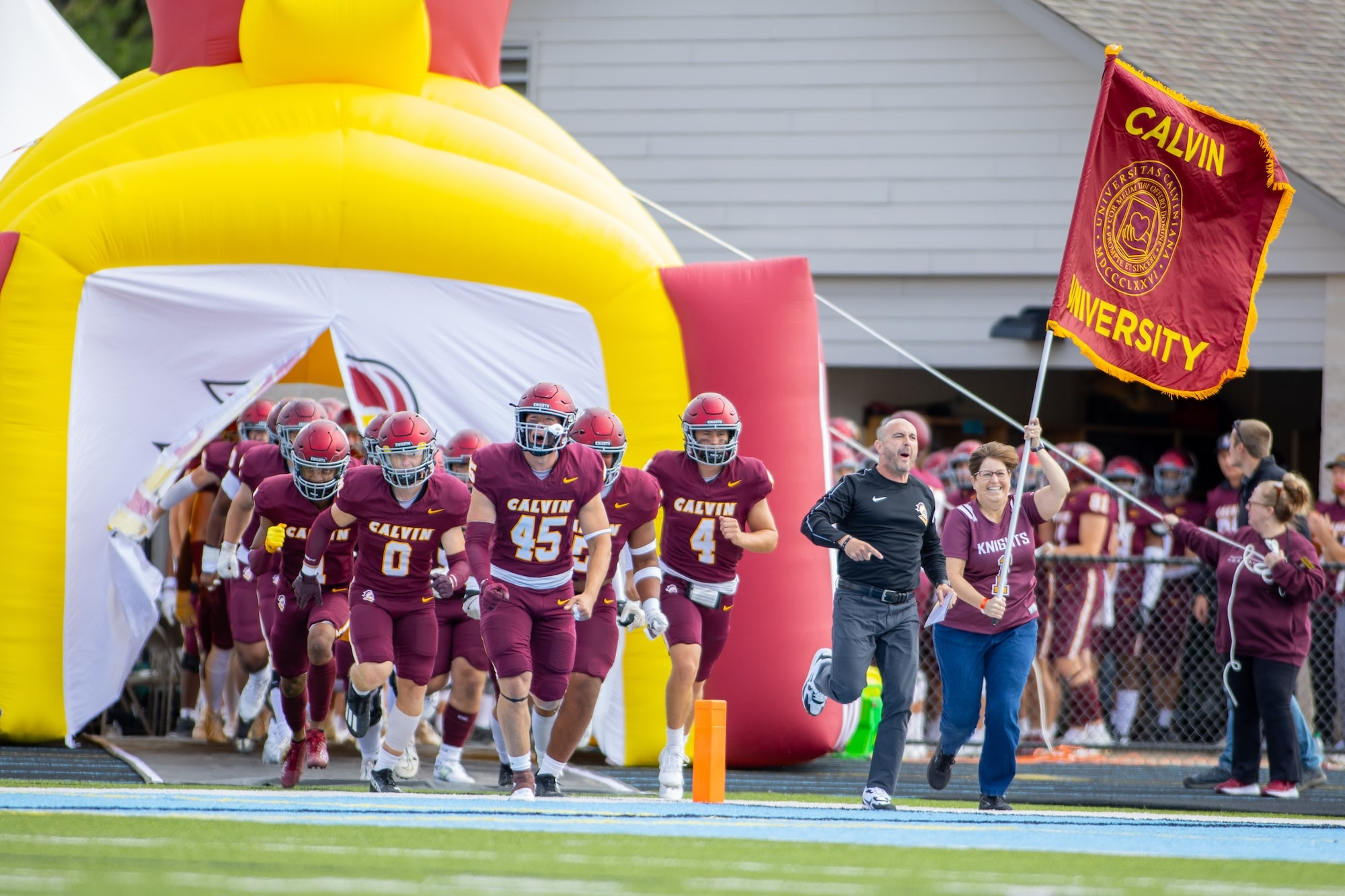 Calvin University football team running out to field, Pastor Mary Hulst running with Calvin flag