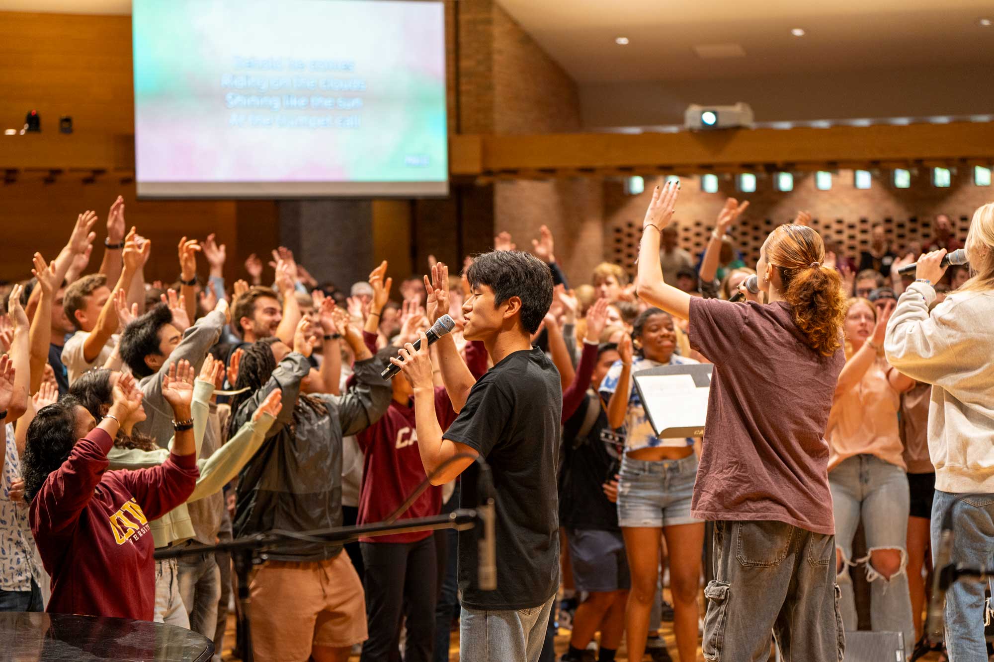 A packed chapel at Calvin University, with students singing with their arms raised, while worship leaders sing into microphones on the stage.