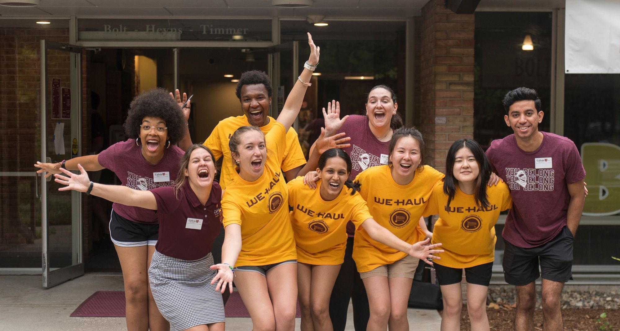 A group of students in maroon and gold "we haul" shirts smile with their arms raised, welcoming new students to campus.