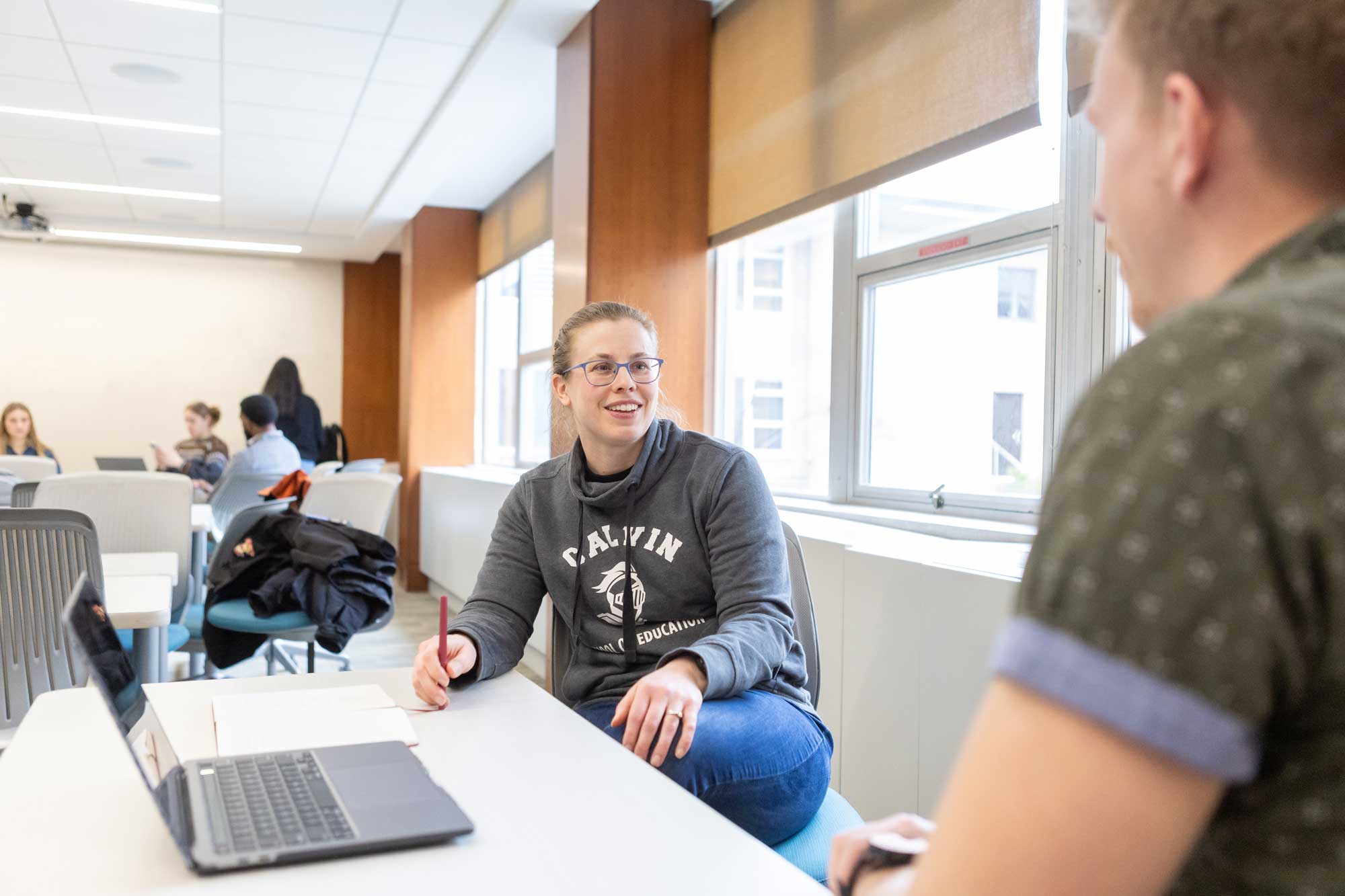 A graduate student in a Calvin School of Education sweatshirt sits at a desk with other students and laptops.