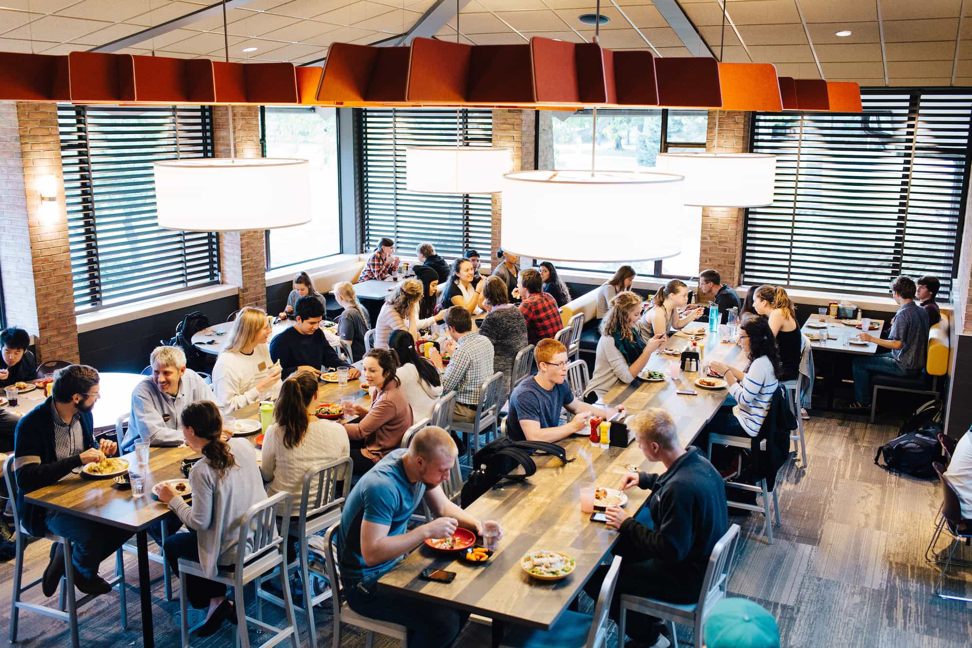 People eat lunch around high-top tables in a dining hall with high ceilings.