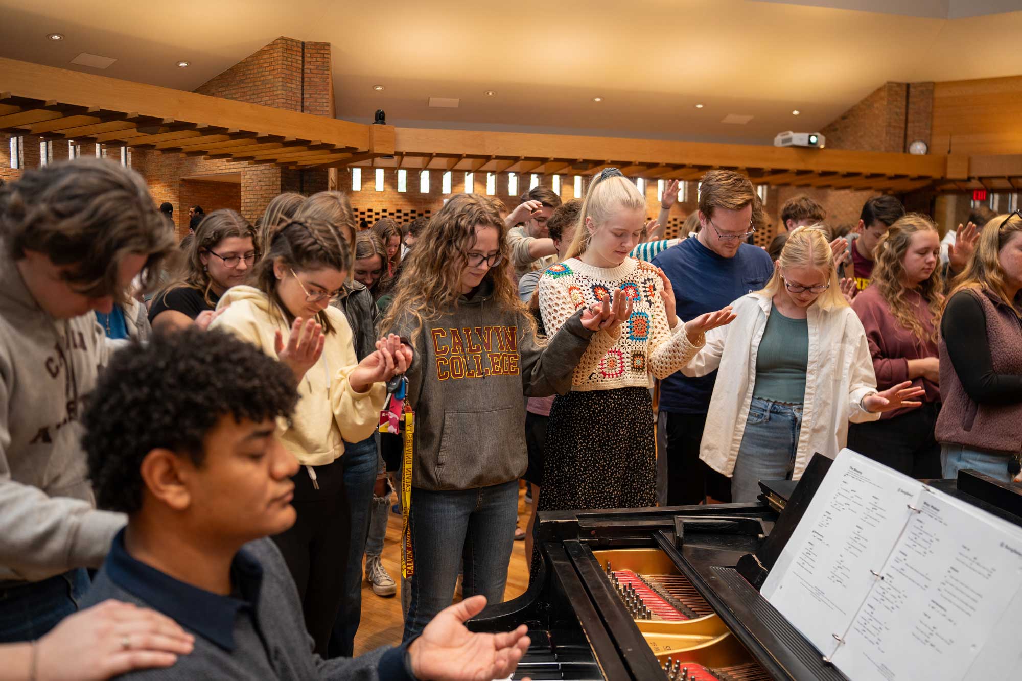Students pray with hands open, gathered around the piano in the Calvin chapel.