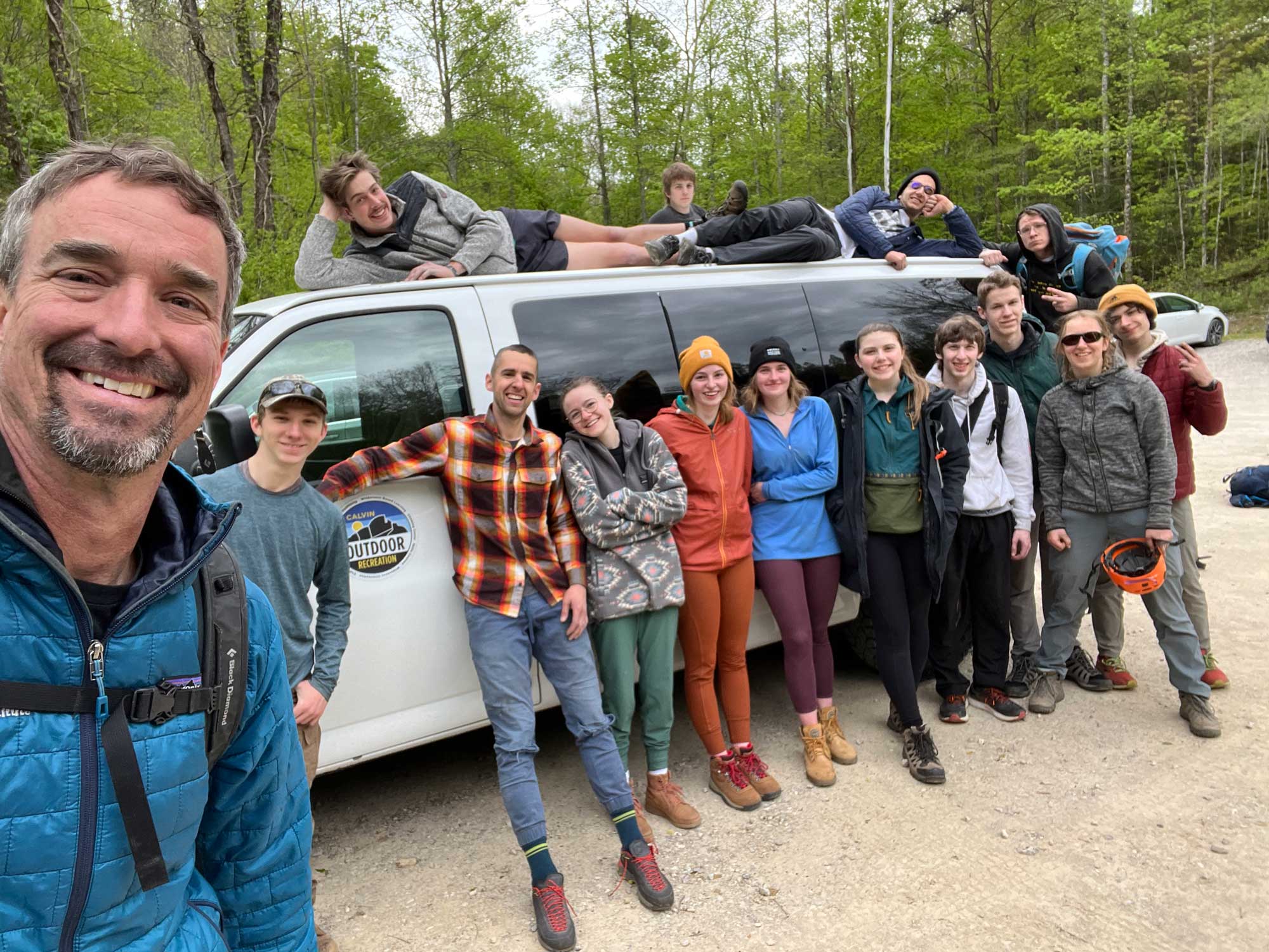 A group of staff and students in front of a 15 passenger van, wearing outdoor gear in the woods.