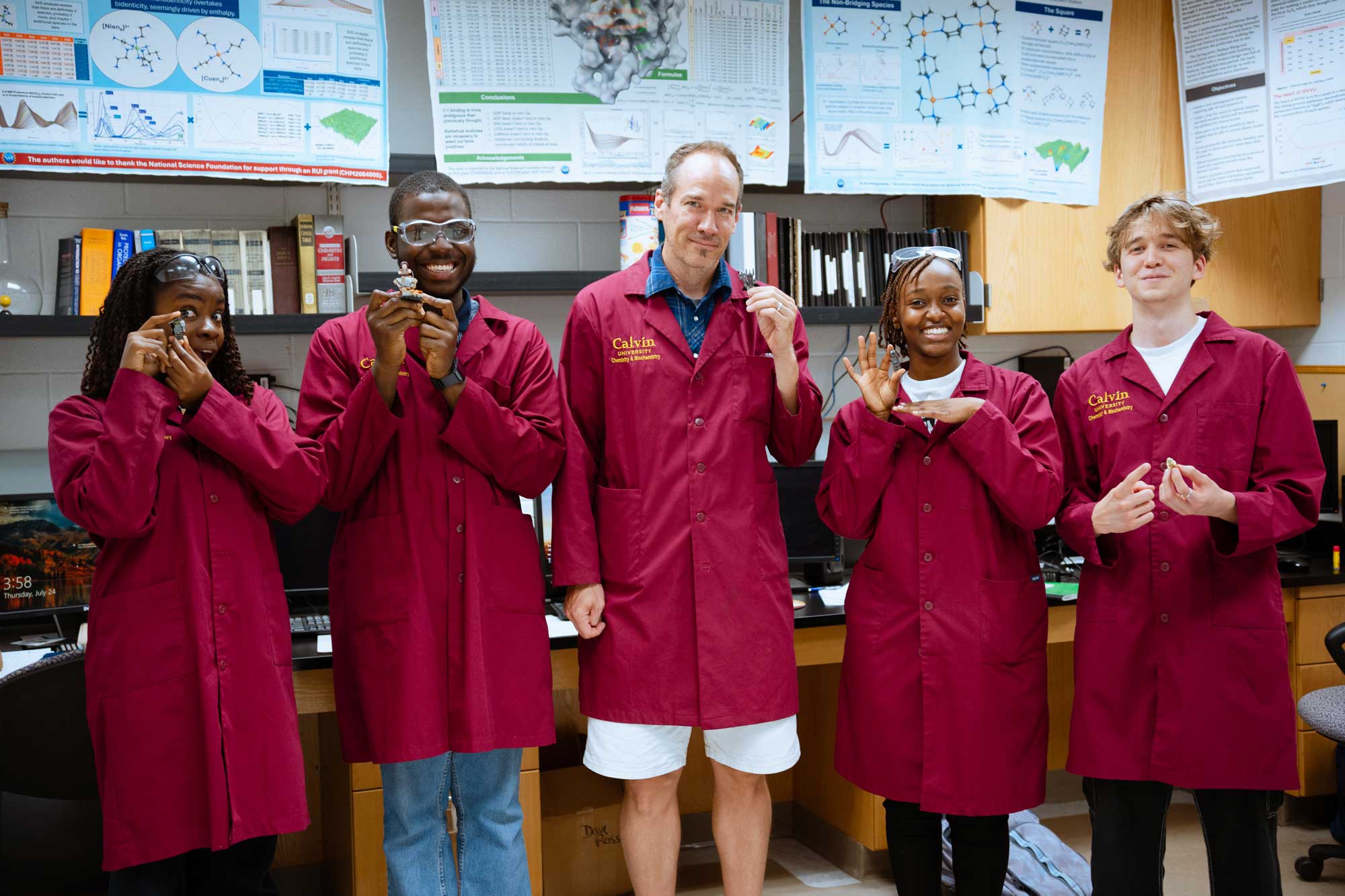 A diverse group of STEM student researchers pose in maroon lab coats with chemistry professor, Doug Vander Griend.