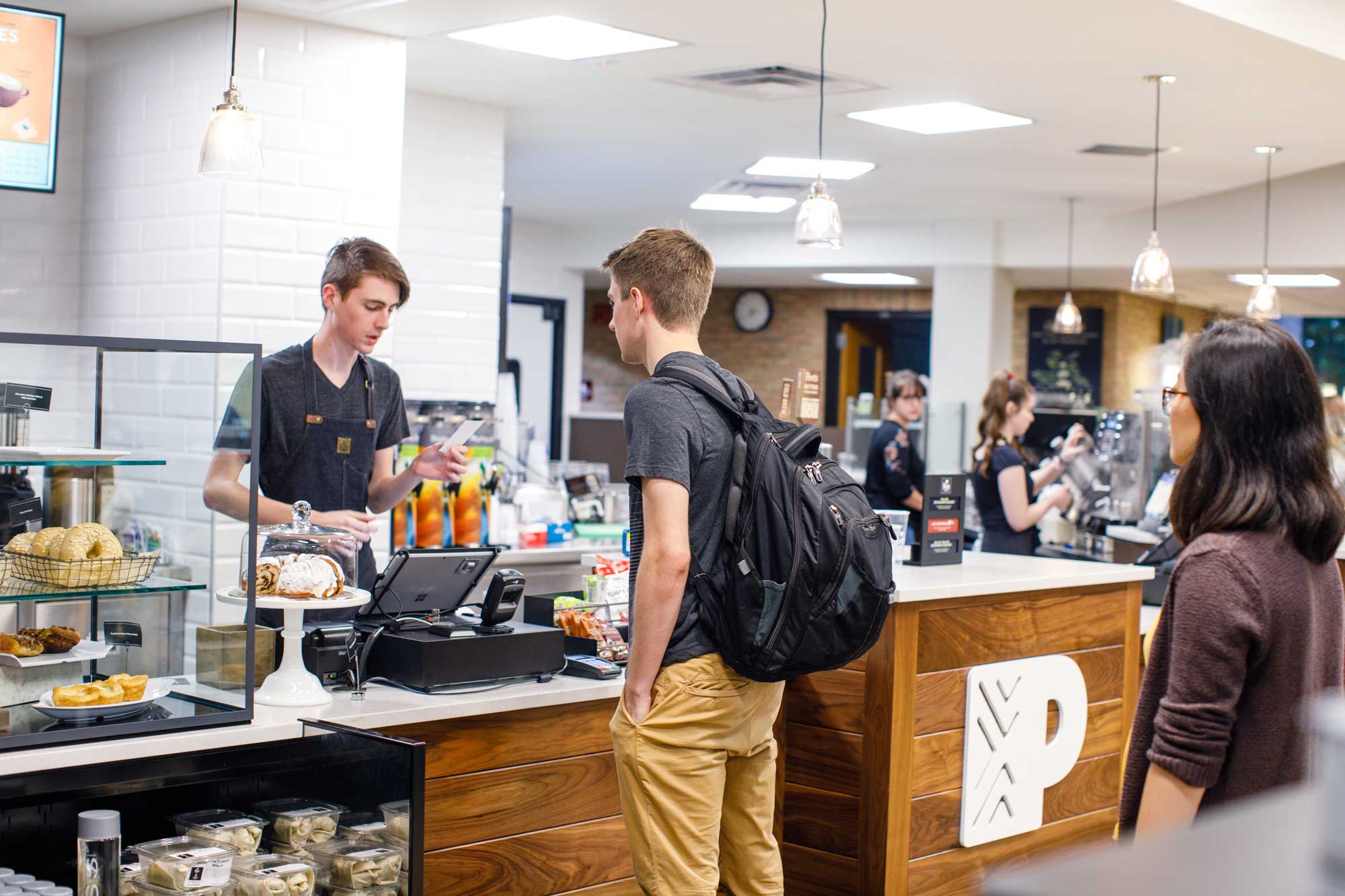 A Peet's Coffee barista takes orders at a counter with a pastry display case, and others making espresso drinks in the background.