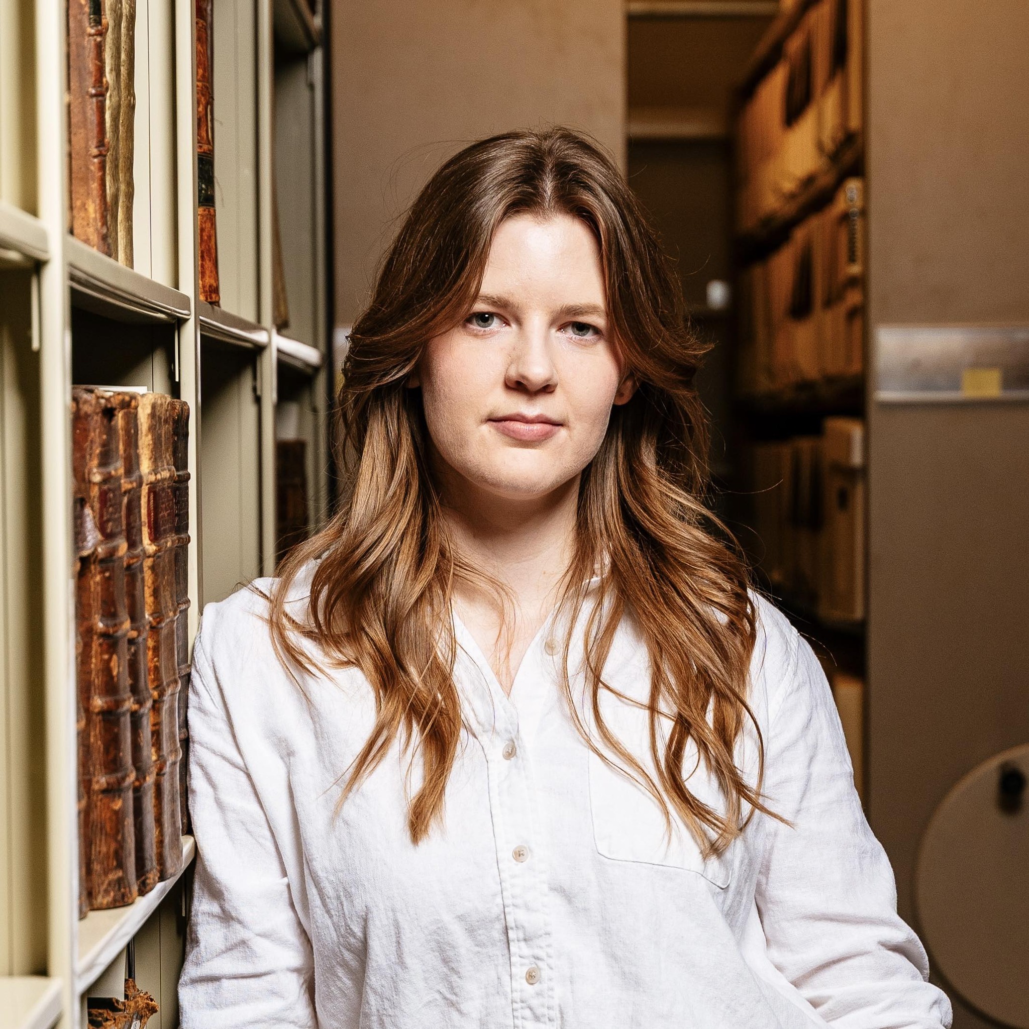 Jen Vos with long caramel brown hair in a white button down shirt leaning against a bookshelf in a library