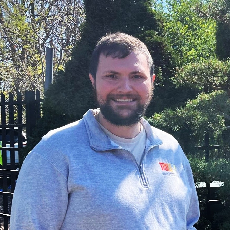 Bryan Burke standing in a light gray pullover sweatshirt with his hands in his pockets in front of pine trees