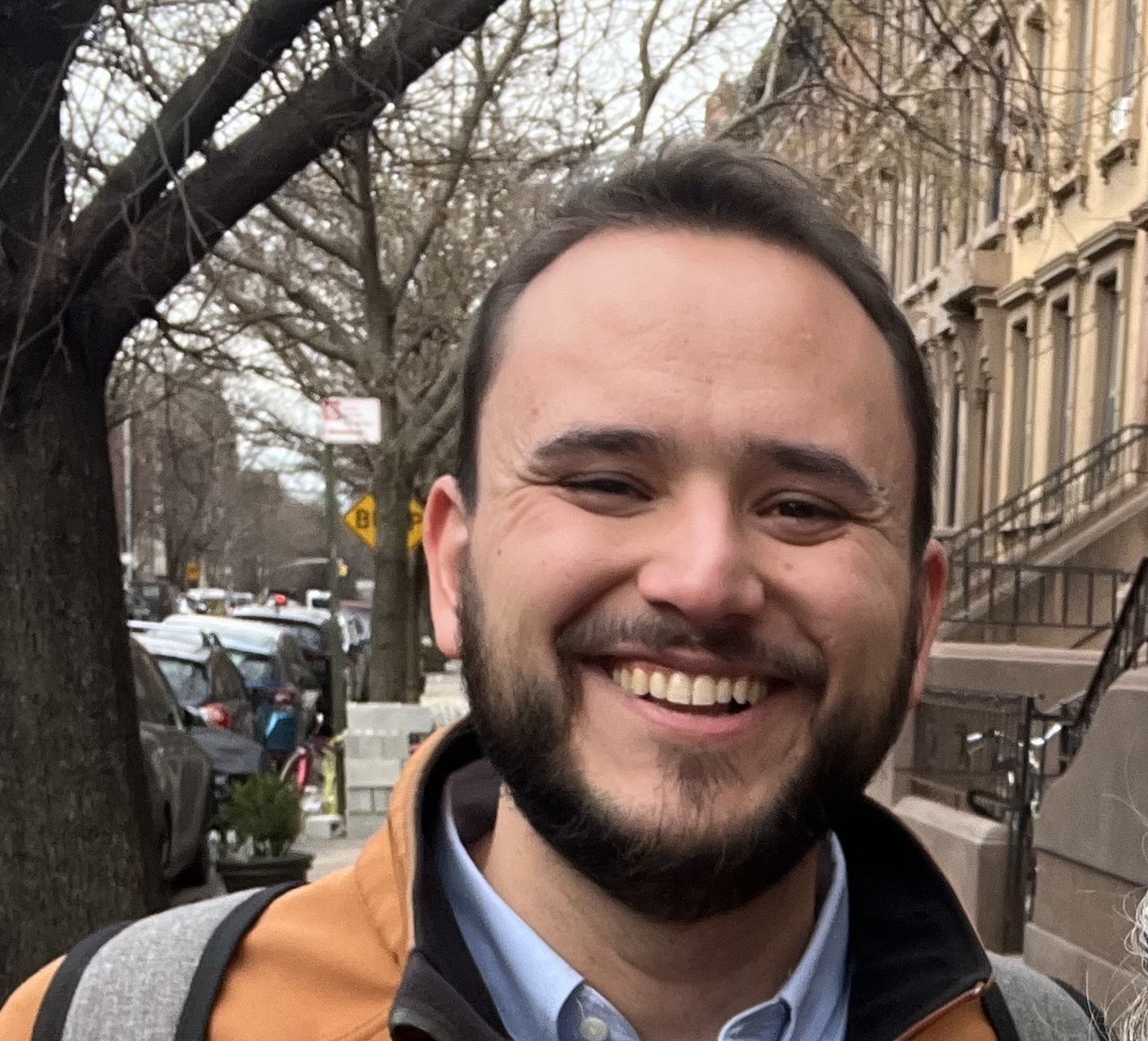 Close up photo of Isaac LaGrand smiling with leafless trees on a city street behind him