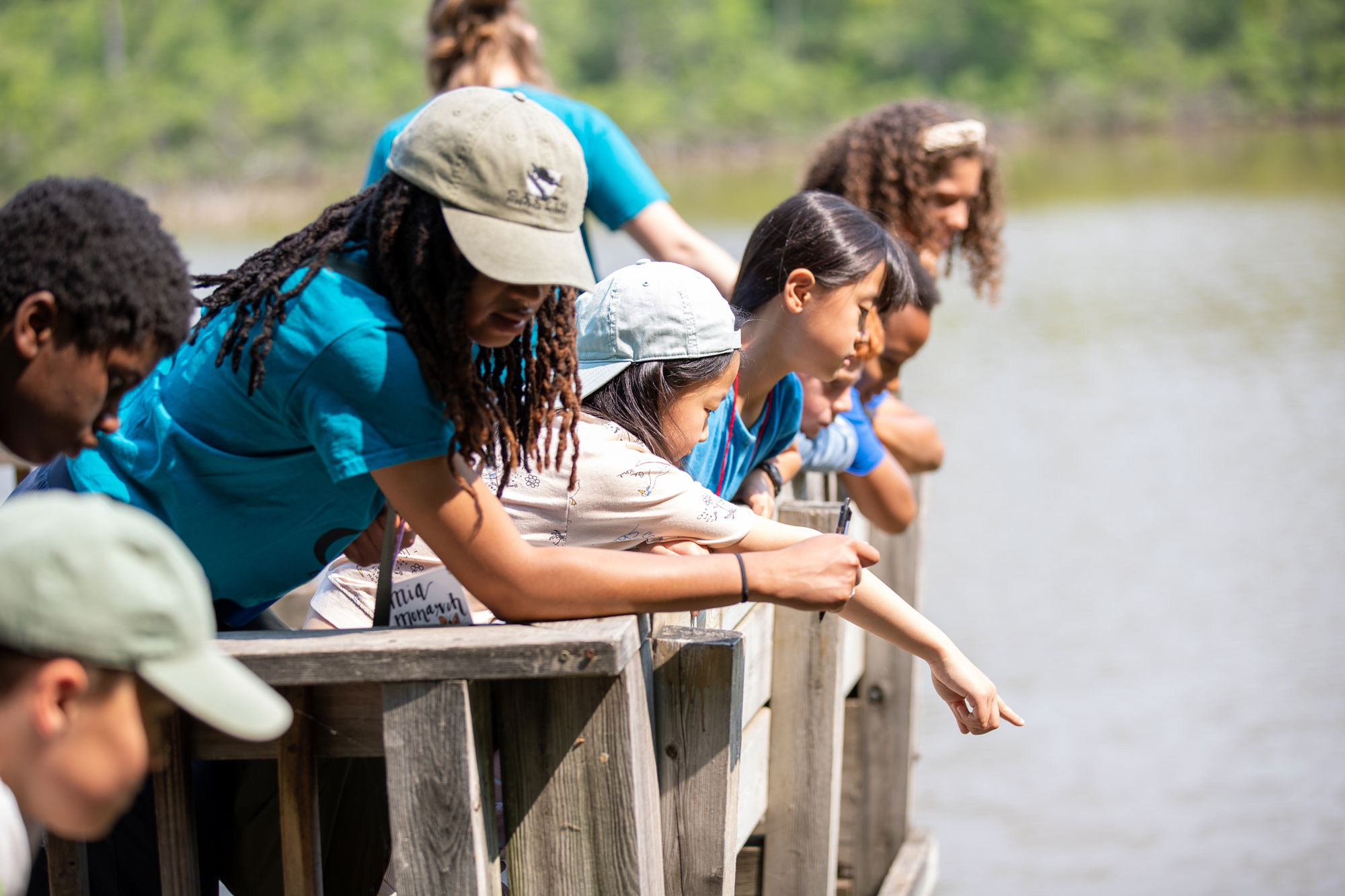 Children and instructors stand on a dock overlooking a pond, pointing to fish.