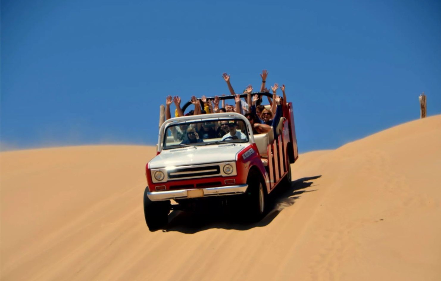 Dunes and a bright blue sky with dune buggy driving downhill with passengers waving their hands in the air