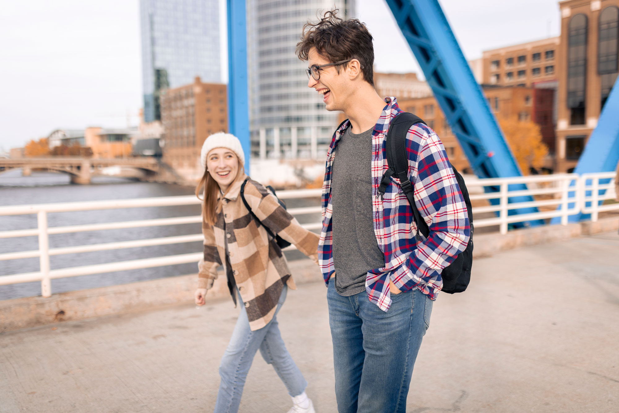 Two students walk across the blue bridge in Grand Rapids, with the riverfront skyline in the background lined with yellow trees.