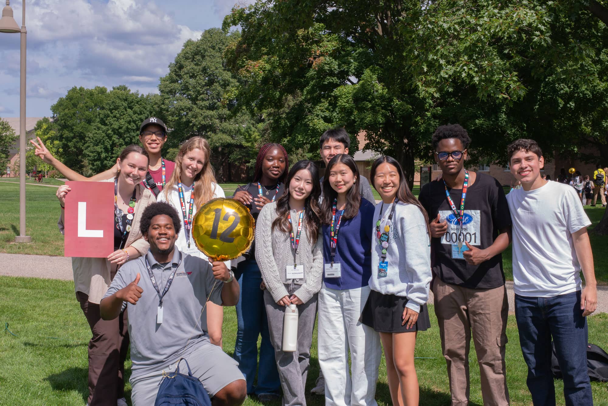 A diverse group of international students smile in a group on Calvin's Commons Lawn, with lanyards and nametags, and a sign and balloon identifying their orientation group.