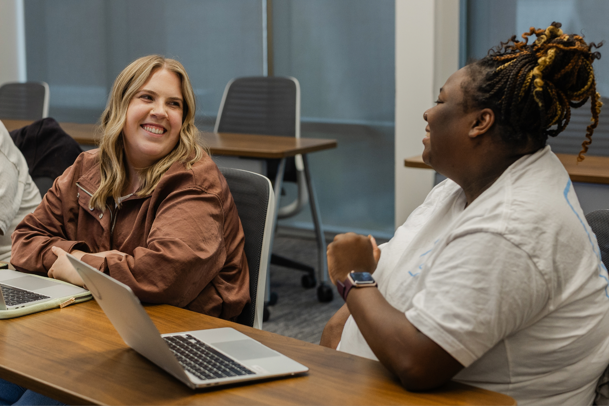 Two female students look at each other and smile. They are in a classroom with laptops open in front of each of them.