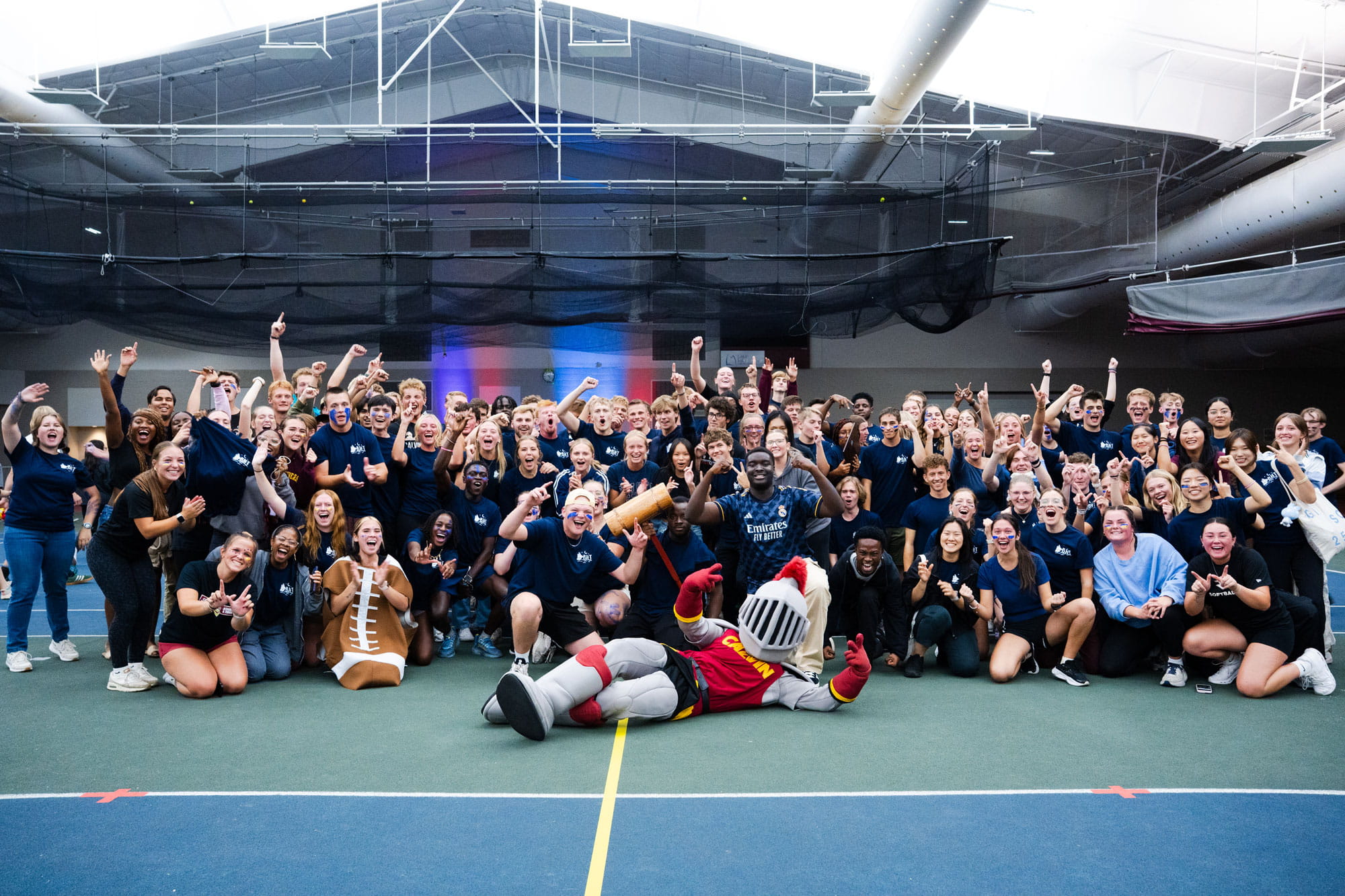 A large group of students with Joust the Knight mascot pose while cheering and holding up number 1, after winning the Chaos Night competition between the Calvin dorms.