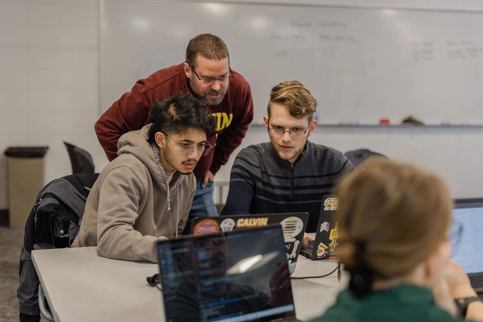 Two computer science students focus on a computer screen while a professor stands near and helps.