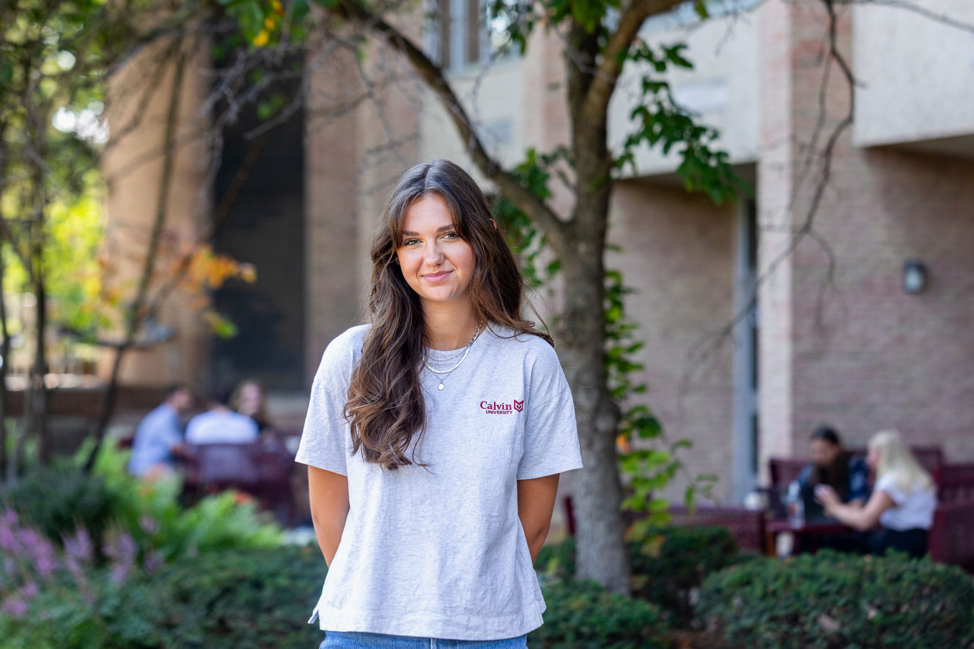 A Calvin student stands in front of the Hekman library while others work at tables behind her.