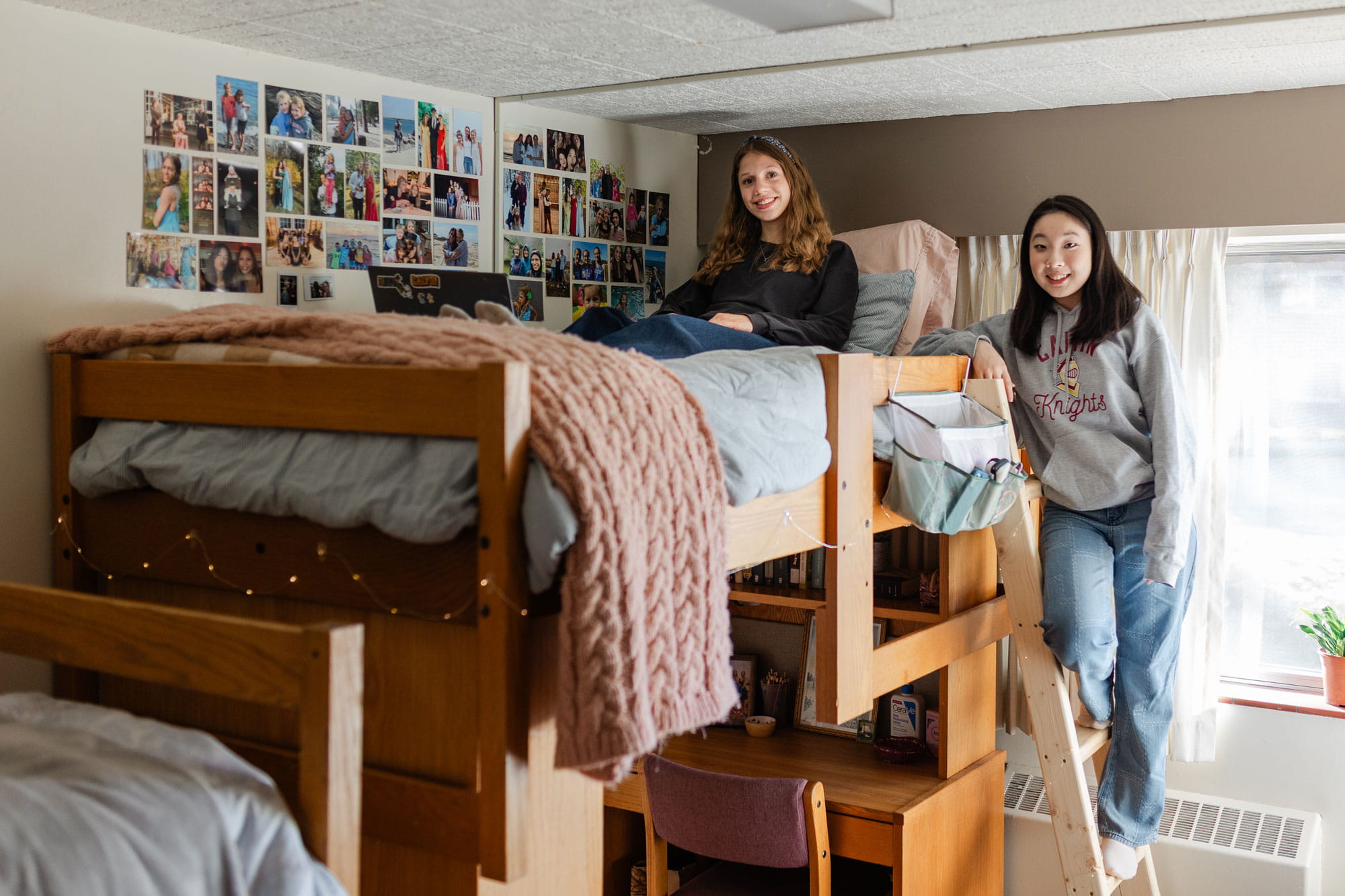 A Calvin student sits on her bed with her laptop and her roommate stands on the bunk bed ladder in their room, showing stacked furniture, a bright window, and photos on the wall.