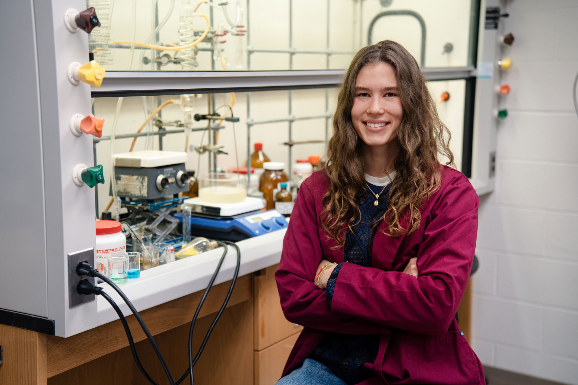 Aerin Baker, a Goldwater Scholar, sits in a research lab wearing a maroon lab coat, smiling confidently into the camera.