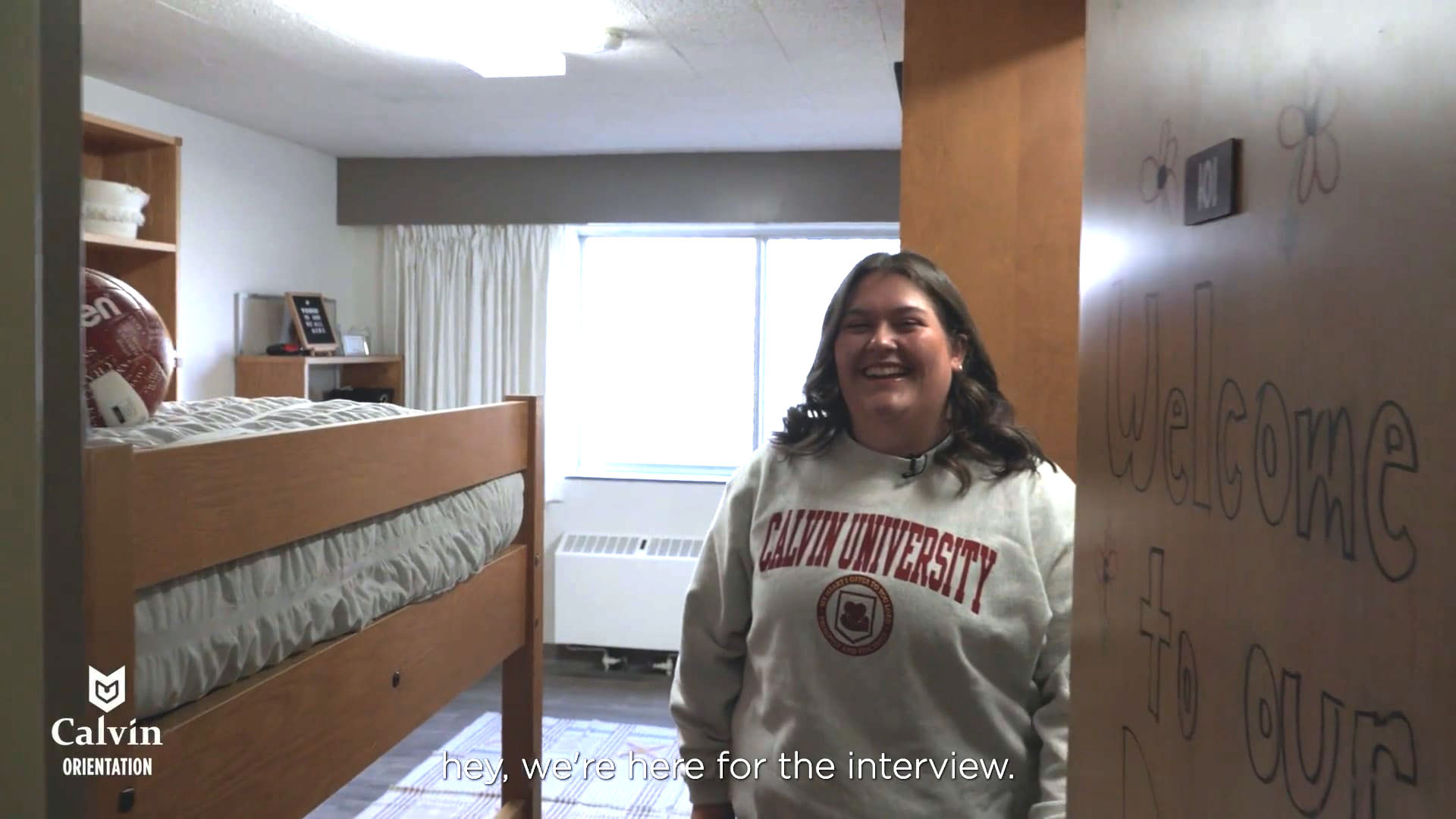 A women opens the door to her dorm and greets the camera.