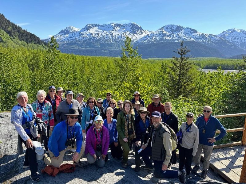 20 people in a group huddle with a green field and snow-dusted mountain in the background