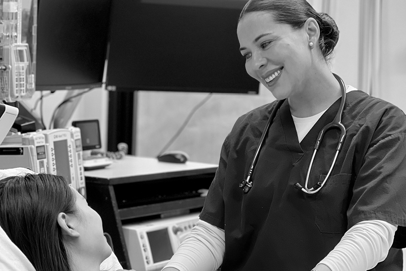 Laura Rivera in scrubs with a stethoscope stands and smiles at the bedside of a patient and her newborn.