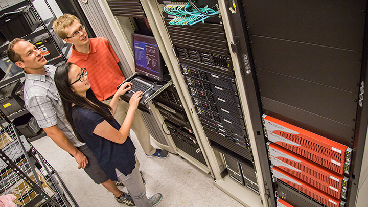 A professor and student watch another student type on a supercomputer terminal's keyboard.