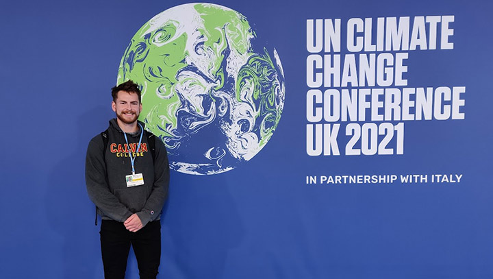 A 2022 male graduate in a Calvin College sweatshirt stands in front of a backdrop with words on it.