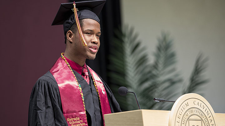 Student in cap and gown standing at a podium provides remarks at formal Commencement ceremony.