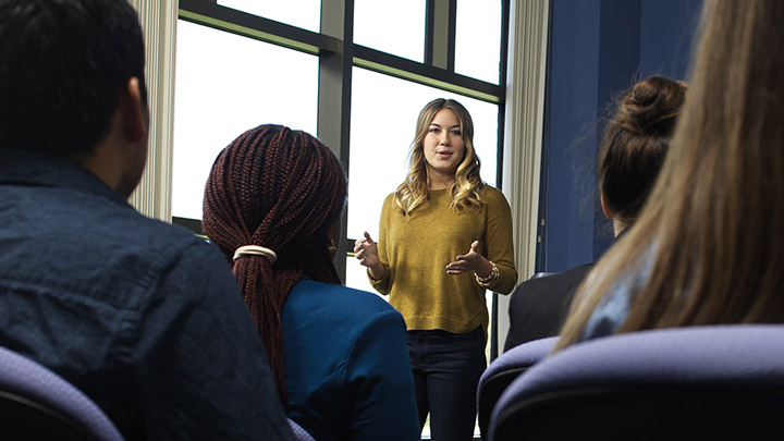 A young woman presents to a classroom of students (view is from a seated student's perspective)