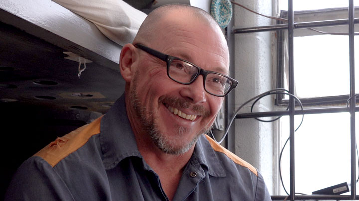 A man with a beaming smile and black-rimmed glasses sits on his bed inside his prison cell.