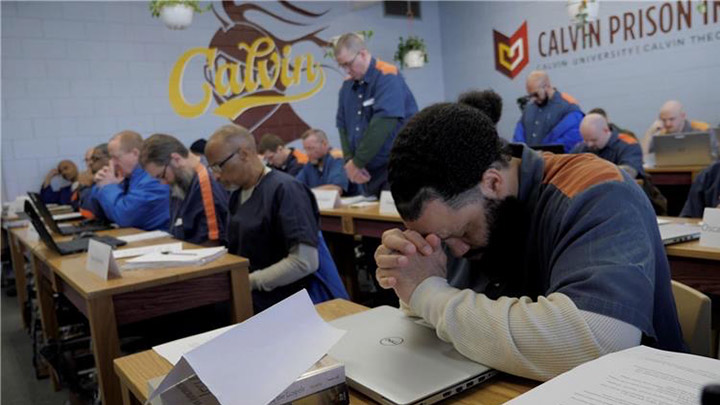 Inmates seated at desks with books and laptops in front of them bow their heads in prayer.