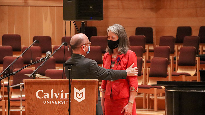 A woman professor stands on stage and is addressed by a man in a suit who is talking to her.