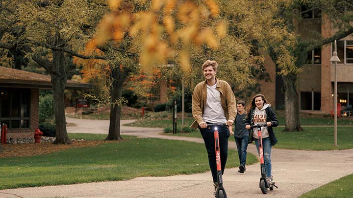 Two students ride electric scooters on a pathway on Calvin University's campus during fall semester.